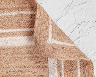 Puede incluir: Una alfombra rectangular tejida con fibras de yute natural en tono bronceado, con una textura trenzada. La alfombra presenta un patrón de bandas anchas de color bronceado alternadas con rayas blancas finas, con una esquina doblada.