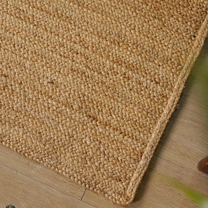 Könnte beinhalten: Nahaufnahme eines rechteckigen, gewebten Jute-Teppichs in Natur-Beige. Der Teppich hat einen geflochtenen Rand und eine strukturierte Oberfläche. Er liegt auf einem hellbraunen Holzboden.