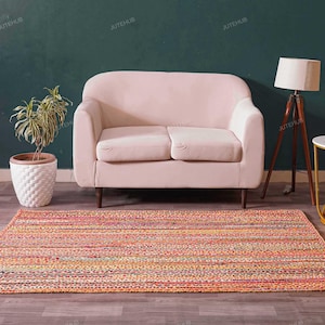 May include: A living room scene featuring a pink loveseat, a colorful striped rug, a wooden tripod lamp, and a small round side table with a marble top. A potted plant adds a touch of greenery.