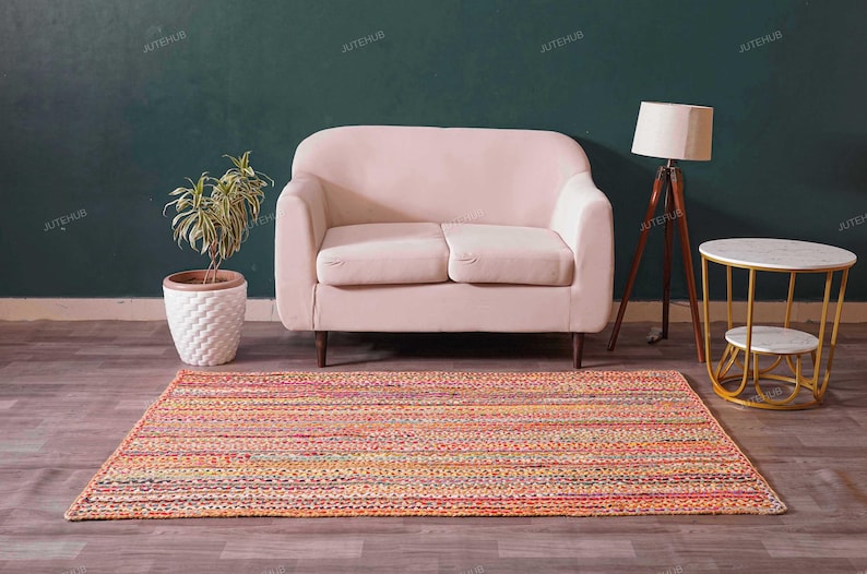 May include: A living room scene featuring a pink loveseat, a colorful striped rug, a wooden tripod lamp, and a small round side table with a marble top. A potted plant adds a touch of greenery.