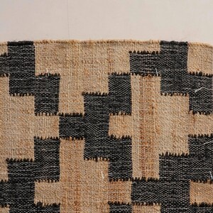 May include: Close-up of a woven rug with a geometric pattern. The rug features a repeating design of black and beige shapes, creating a textured appearance. The edge of the rug has a fringe detail.