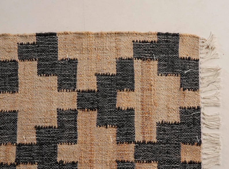 May include: Close-up of a woven rug with a geometric pattern. The rug features a repeating design of black and beige shapes, creating a textured appearance. The edge of the rug has a fringe detail.
