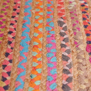 May include: Close-up of a colorful braided jute rug. The rug features vertical stripes of natural jute interspersed with vibrant braids of pink, blue, orange, and gray fabric. The texture is coarse and the colors are bright.