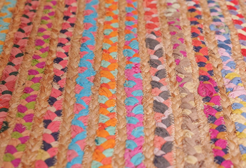 May include: Close-up of a colorful braided jute rug. The rug features vertical stripes of natural jute interspersed with vibrant braids of pink, blue, orange, and gray fabric. The texture is coarse and the colors are bright.