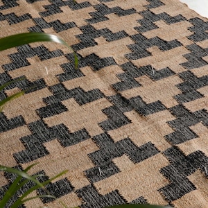 May include: A woven rug with a geometric pattern in black and beige. The rug's design features a repeating pattern of interlocking shapes, creating a textured surface. The rug is made of natural fibres, and the colour palette is neutral.