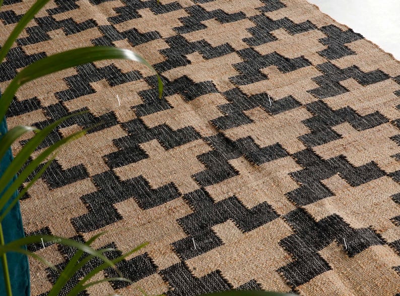 May include: A woven rug with a geometric pattern in black and beige. The rug's design features a repeating pattern of interlocking shapes, creating a textured surface. The rug is made of natural fibres, and the colour palette is neutral.