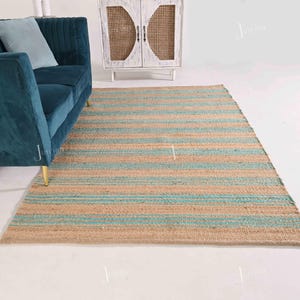 Op de afbeelding: Een rechthoekig jute vloerkleed met horizontale strepen in natuurlijk beige en lichtblauw. Het vloerkleed ligt op een witte vloer, met een turquoise bank en een witte kast op de achtergrond. Het ontwerp van het vloerkleed voegt een vleugje kuststijl toe.