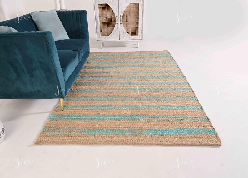 Op de afbeelding: Een rechthoekig jute vloerkleed met horizontale strepen in natuurlijk beige en lichtblauw. Het vloerkleed ligt op een witte vloer, met een turquoise bank en een witte kast op de achtergrond. Het ontwerp van het vloerkleed voegt een vleugje kuststijl toe.
