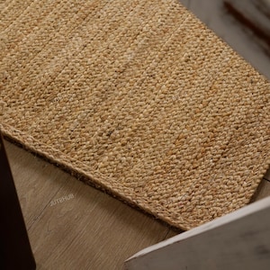 May include: A rectangular jute rug with a woven texture. The rug is a natural tan color and appears to be made of thick, durable fibers. The rug is placed on a light-colored wooden floor, with a portion of a chair visible.