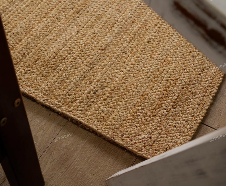 May include: A rectangular jute rug with a woven texture. The rug is a natural tan color and appears to be made of thick, durable fibers. The rug is placed on a light-colored wooden floor, with a portion of a chair visible.