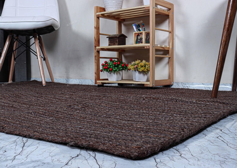 Op de afbeelding: Een rechthoekig vloerkleed met een dikke, gevlochten jute textuur in donkerbruin. Het kleed ligt plat op een lichte vloer met een marmerachtig patroon, voor een houten rek en een witte stoel met houten poten.