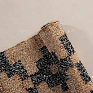 May include: A rolled-up rug with a geometric pattern in natural tan and black. The rug's design features a repeating zigzag pattern. The rug is made of a woven material, possibly jute or a similar fibre.