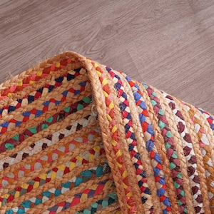 May include: Close-up of a braided jute rug with a mix of colorful threads. The rug features a natural tan color with interwoven sections of red, blue, yellow, and green. The edges are neatly finished with a thick, braided border.