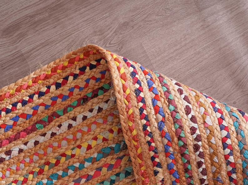 May include: Close-up of a braided jute rug with a mix of colorful threads. The rug features a natural tan color with interwoven sections of red, blue, yellow, and green. The edges are neatly finished with a thick, braided border.