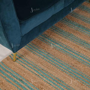 Op de afbeelding: Een teal fluwelen bank met gouden poten staat op een natuurlijk jute vloerkleed. Het vloerkleed heeft een gestreept patroon in bruin en teal. De hoek van de bank is zichtbaar en toont de zachte textuur en het moderne design.