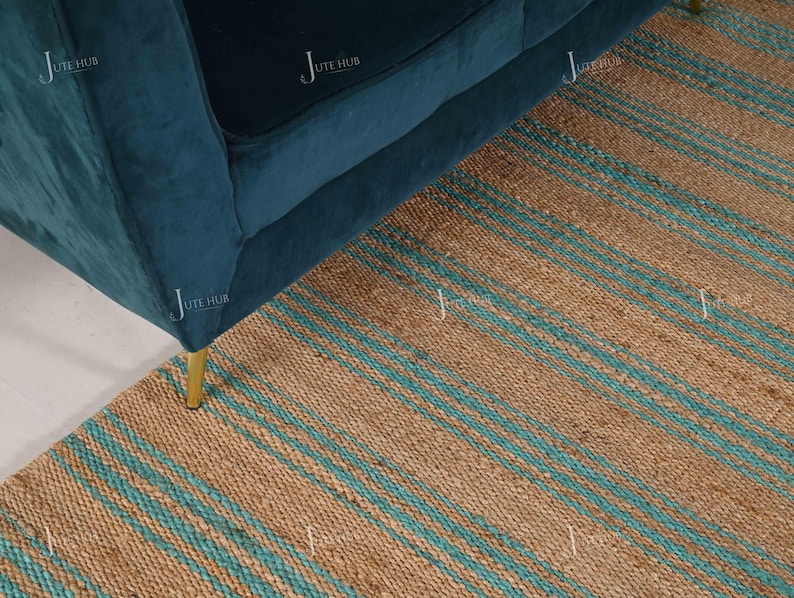 Op de afbeelding: Een teal fluwelen bank met gouden poten staat op een natuurlijk jute vloerkleed. Het vloerkleed heeft een gestreept patroon in bruin en teal. De hoek van de bank is zichtbaar en toont de zachte textuur en het moderne design.
