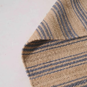 Könnte beinhalten: Ausschnitt eines gewebten Teppichs mit natürlichem beigefarbenem Hintergrund und gleichmäßig verteilten blauen Streifen. Die Textur des Teppichs ist sichtbar, ein Teil ist umgeschlagen.