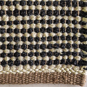 May include: A woven rug featuring a repeating pattern of alternating black and cream-colored horizontal rows. The texture is coarse and fibrous, with a solid brown border along the bottom edge. The weave is tight and uniform throughout the surface.