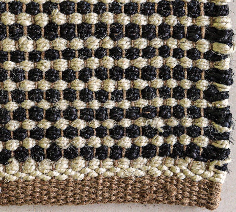 May include: A woven rug featuring a repeating pattern of alternating black and cream-colored horizontal rows. The texture is coarse and fibrous, with a solid brown border along the bottom edge. The weave is tight and uniform throughout the surface.