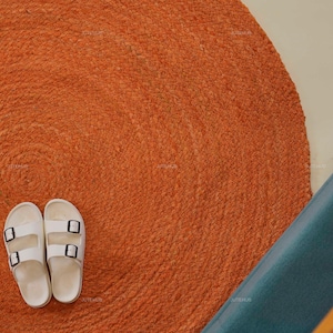 Könnte beinhalten: Draufsicht auf einen runden, orangefarbenen Jute-Teppich mit weißen Sandalen. Die Sandalen haben schwarze Schnallen. Ein türkisfarbener Untergrund und ein Teil einer Holzkonstruktion sind im Hintergrund zu sehen.