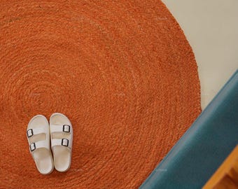 Puede incluir: Vista aérea de una alfombra redonda de yute naranja con sandalias blancas. Las sandalias tienen hebillas negras. Un fondo azul turquesa y una sección de una estructura de madera están en el fondo.