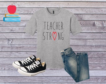 May include: A gray t-shirt with the text "TEACHER STRONG" and a red heart. The shirt is paired with blue jeans and black sneakers.