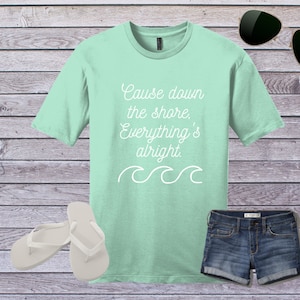 May include: A light green t-shirt with the text "Cause down the shore, Everything's alright." in white. The shirt has a white wave design below the text. The shirt is displayed on a wooden background with a pair of white flip flops, a pair of sunglasses, and a pair of blue denim shorts.