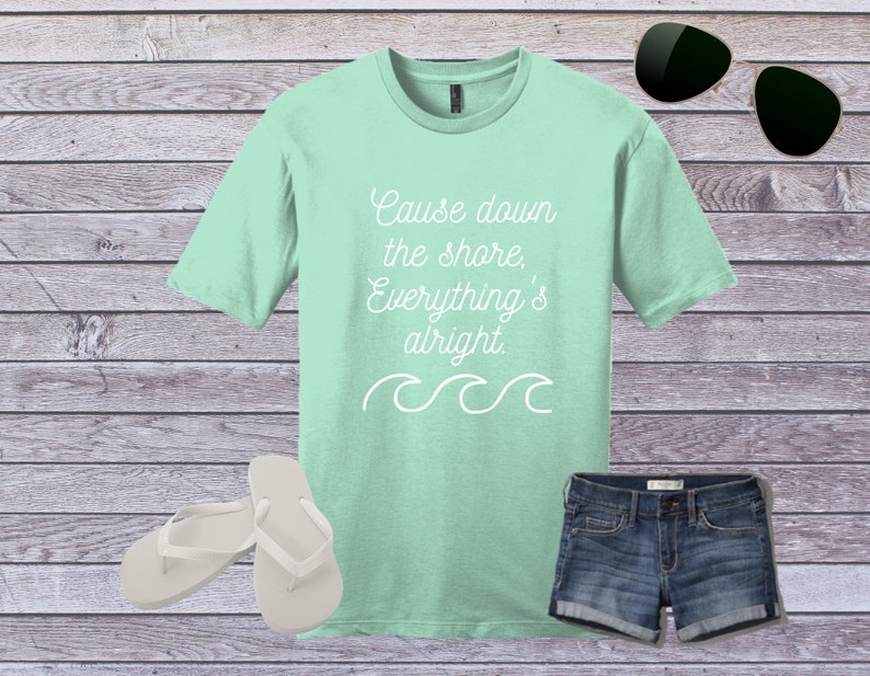 May include: A light green t-shirt with the text "Cause down the shore, Everything's alright." in white. The shirt has a white wave design below the text. The shirt is displayed on a wooden background with a pair of white flip flops, a pair of sunglasses, and a pair of blue denim shorts.