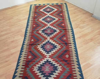 May include: A long, narrow rug with a geometric pattern in shades of red, blue, green, and beige. The rug has a fringe on both ends.