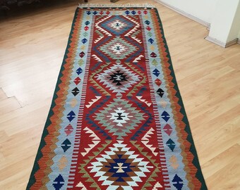 May include: A long, narrow rug with a traditional geometric pattern in shades of red, blue, green, and brown. The rug has a fringe on both ends.