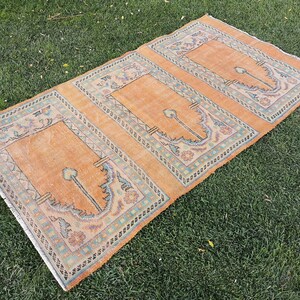 Puede incluir: Tres alfombras de oración de color naranja con intrincados diseños florales azules y blancos. Las alfombras están dispuestas sobre una superficie de hierba verde.