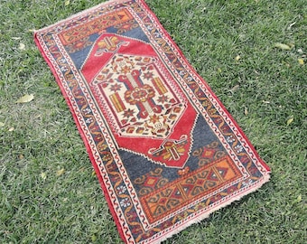 May include: A red, blue, and orange patterned rug with a traditional design. The rug has a fringed edge and is laid out on a green grassy surface.
