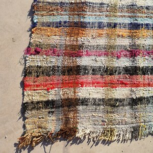 May include: A colorful, handwoven rug with a striped pattern in shades of red, orange, yellow, blue, green, black, and white. The rug has a frayed edge and is made of recycled materials.