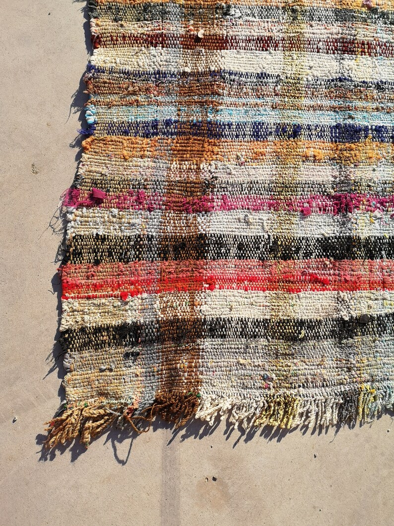 May include: A colorful, handwoven rug with a striped pattern in shades of red, orange, yellow, blue, green, black, and white. The rug has a frayed edge and is made of recycled materials.