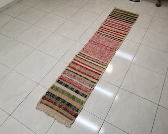 May include: A long, narrow, multi-colored striped rug with fringe on the ends. The rug is made of a woven fabric and has a variety of colors, including red, green, black, and white.