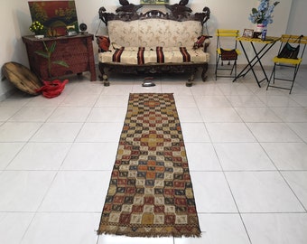 May include: A long, rectangular rug with a geometric pattern in shades of brown, red, yellow, and blue. The rug is laid out on a white tiled floor in a room with furniture.