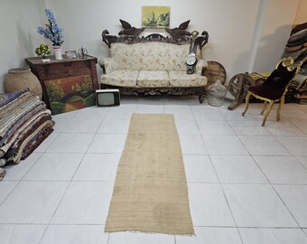 May include: A rectangular, beige rug with horizontal stripes is centered on a tiled floor. The rug is in a room with antique furniture, including a carved wooden sofa, a wooden table, and a decorative chair. Other items include a painting, a vintage TV, and a large glass bottle.
