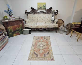 May include: A vintage rug with a cream background and a central orange and pink design. The rug is laid out on a tiled floor in a room with a couch and other furniture.