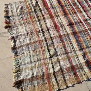 May include: A handwoven rug with a striped pattern in shades of brown, beige, red, and blue. The rug has a fringe border and is made of recycled materials.