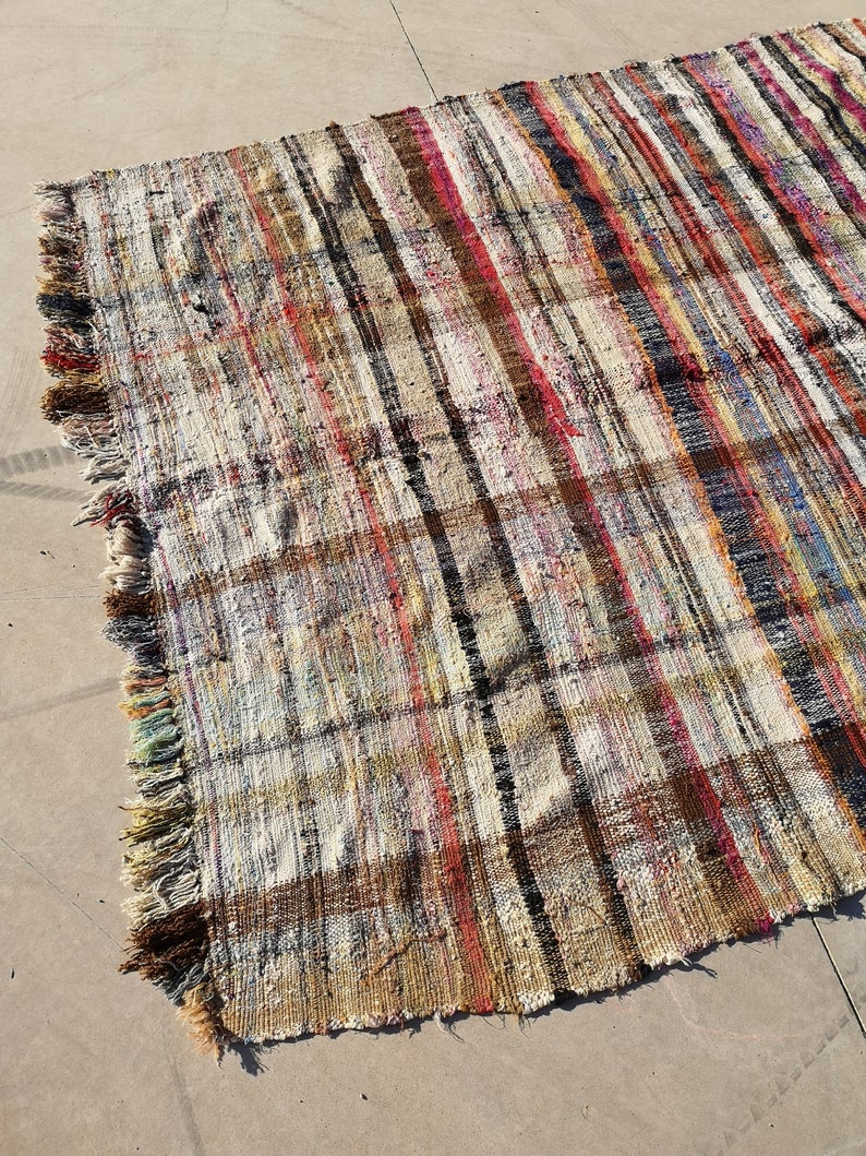 May include: A handwoven rug with a striped pattern in shades of brown, beige, red, and blue. The rug has a fringe border and is made of recycled materials.