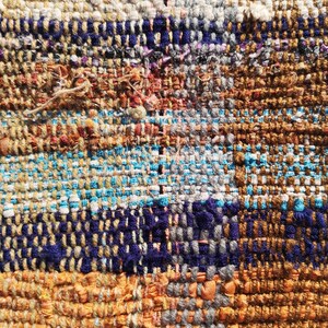 May include: Close-up of a woven tapestry with a variety of colors, including brown, blue, white, and gray. The tapestry is made of different types of yarn and has a textured surface.