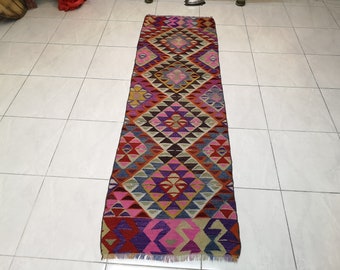 May include: A long, colorful rug with a geometric pattern in shades of pink, purple, blue, red, and brown. The rug has a fringe border.