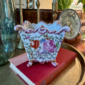 May include: A white and floral porcelain planter with a scalloped rim, gold accents, and small feet. The planter features a vibrant design of roses and other flowers. It rests on top of a red book.