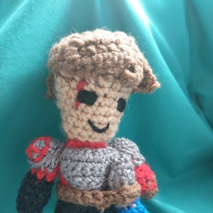 Pode incluir: Uma figura de brinquedo de crochê que representa uma pessoa com armadura cinza e um cinto marrom, segurando uma espada azul. A figura tem cabelo castanho e uma cicatriz vermelha no rosto.