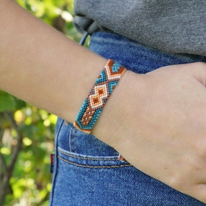 May include: A woven friendship bracelet featuring a geometric pattern in shades of teal, burnt orange, white, and brown. The bracelet is constructed from multiple strands of thread intricately knotted together to form a repeating diamond design.