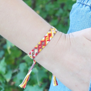 May include: A woven friendship bracelet featuring a repeating geometric chevron pattern in vibrant shades of red, yellow, white, and brown. The bracelet is finished with thin, knotted braided ties at each end, showcasing intricate thread work.