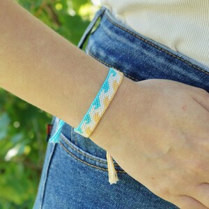 May include: A woven friendship bracelet featuring a repeating chevron pattern in alternating shades of light blue, white, and soft yellow. The bracelet is constructed from thin thread and includes dangling thread ends at the bottom edge.
