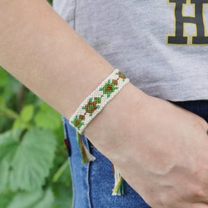 May include: A woven friendship bracelet with a cream-coloured base and a repeating pattern of green and brown diamond shapes. The bracelet is tied with green, brown, and cream-coloured threads. The bracelet is approximately 1.3 cm wide.