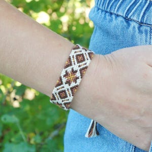 May include: A woven friendship bracelet in shades of brown, tan, and white, with a geometric diamond pattern. The bracelet is worn on a wrist, with a denim background. The bracelet has a tassel at the end.