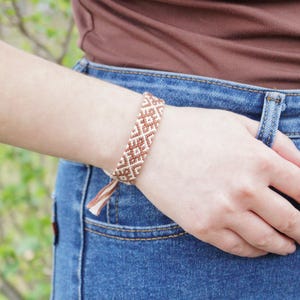 Könnte beinhalten: Ein gewebtes Freundschaftsarmband in Braun- und Cremetönen mit einem geometrischen Muster wird am Handgelenk getragen. Die Person trägt ein braunes Oberteil und eine blaue Jeans. Das Armband ist traditionell.